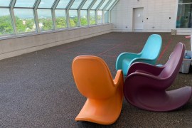 Three brightly colored, modern-style plastic rocking chairs are in a large room. The walls are lined with side-to-side windows, which fill the room with natural light.