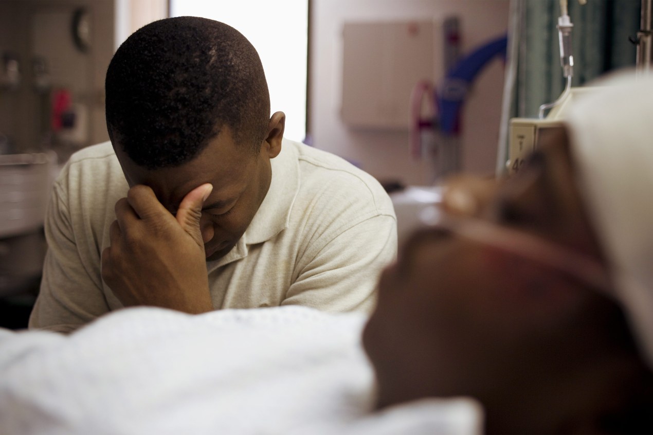 A photo of a Black man sitting beside his son in a hospital bed, covering his face.